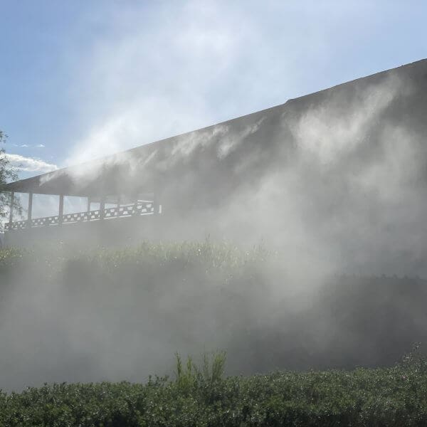 Misting system installation in a residential setting, showcasing water misting against a backdrop of a house and greenery, emphasizing cooling and comfort.